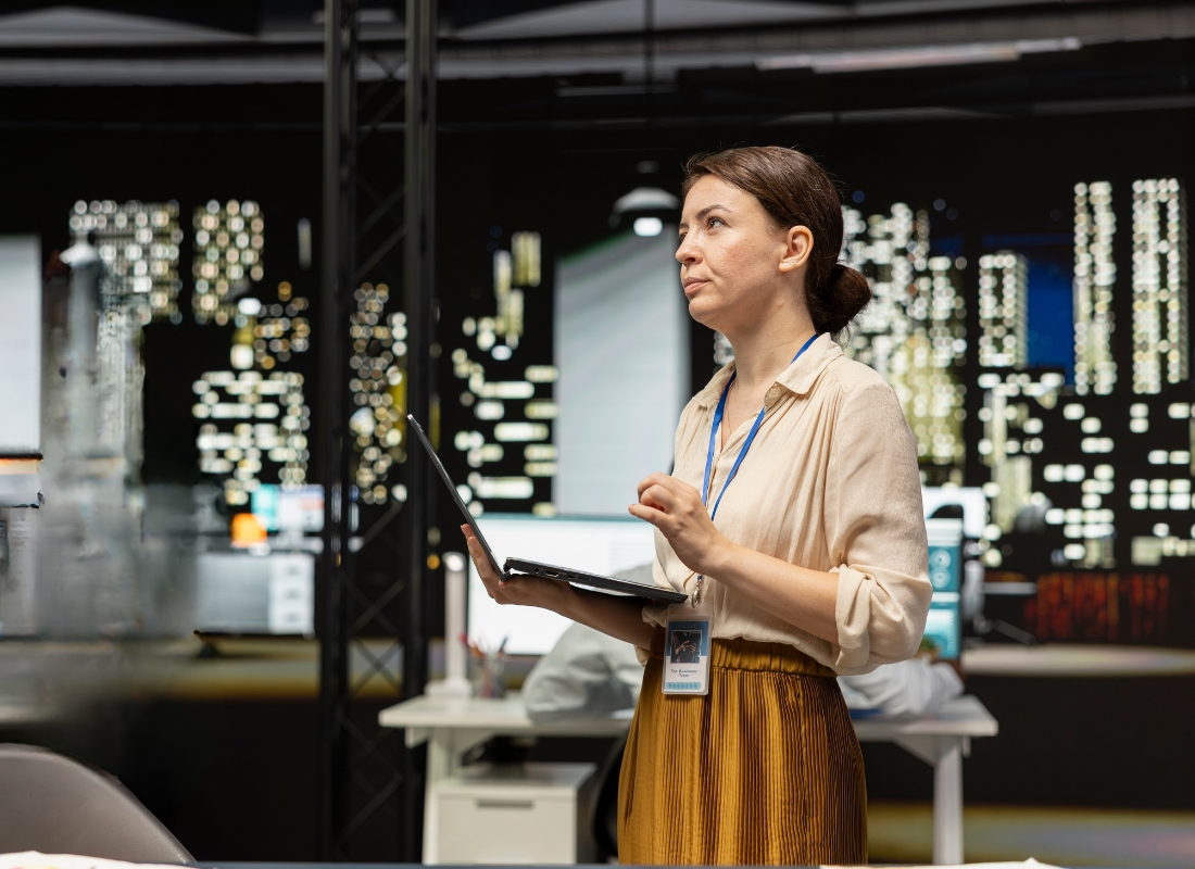 prikaz žene koja stoji u firmi, drži laptop u ruci i zamišljeno gleda u stranu