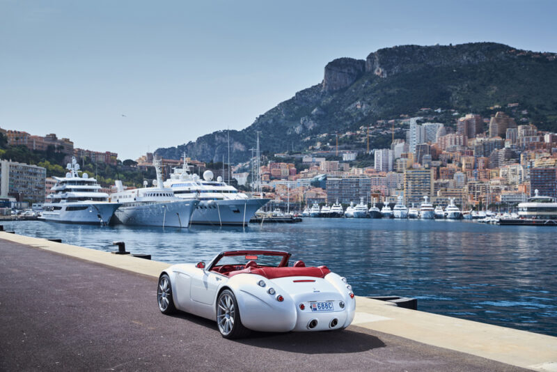 Beli sportski kabriolet parkiran pored marine u Monaku sa luksuznim jahtama i planinom u pozadini