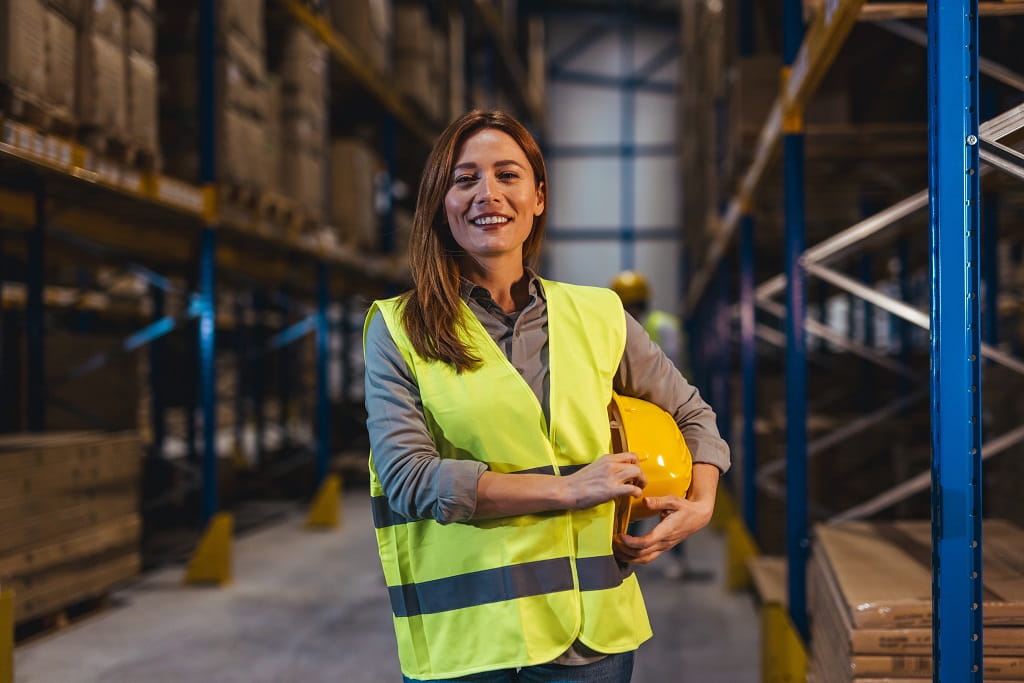 Radnica u magacinu sa reflektujućim prslukom i zaštitnom kacigom među policama sa robom