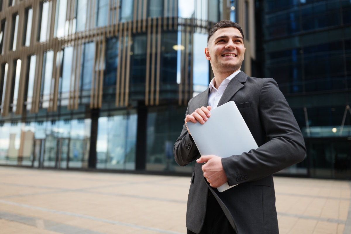 dečko u odelu stoji ispred zgrade sa laptopom u ruci i smeje se